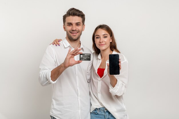 Young happy couple in white shirts hold in hand credit bank card using smartphone isolated on white