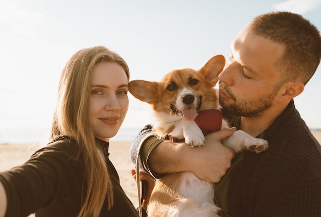 犬と若い幸せなカップルは、ビーチでselfieを取る。美しい少女と男とコーギーの子犬