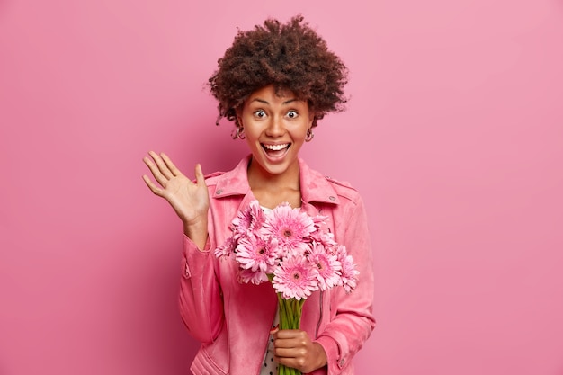 Young happy curly haired Afro American lady recieves beautiful bouquet of gerberas for spring holiday keeps palm raised wears stylish jacket isolated over pink studio wall