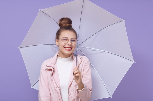 라일락 배경 위에 고립 된 하얀 우산, 옅은 분홍색 재킷과 흰색 셔츠를 입고 매듭을 가진 아가씨, 행복을 표현하는 젊은 행복 감정 소녀.