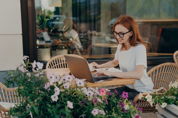 Молодая счастливая европейская женщина, сидящая за столиком в кафе на открытом воздухе с ноутбуком и чашкой кофе, улыбающаяся рыжая женщина в очках, наслаждающаяся удаленной работой в кафе или обучающаяся онлайн