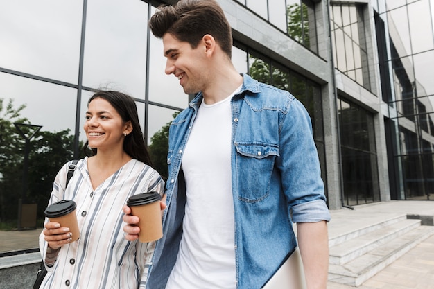 молодые счастливые возбужденные удивительные любящие студенты пары на открытом воздухе на улице, гуляя, пьют кофе, разговаривают друг с другом.