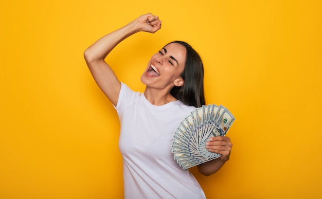 Молодая счастливая возбужденная красивая женщина эмоционально смотрит в камеру с кучей денег в руках и развлекается на желтом фоне