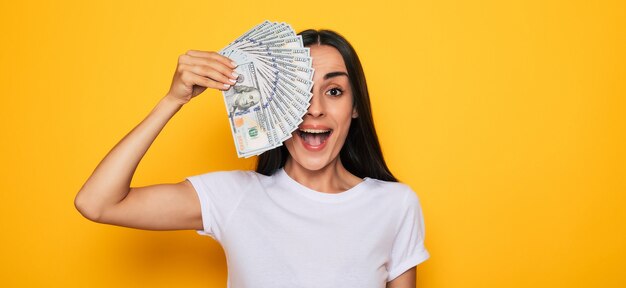 Young happy excited beautiful woman emotionally looking in the camera with a lot of money in her hands and having fun isolated on yellow background