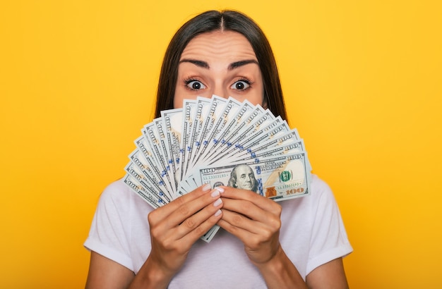 Young happy excited beautiful woman emotionally looking in the camera with a lot of money in her hands and having fun isolated on yellow background