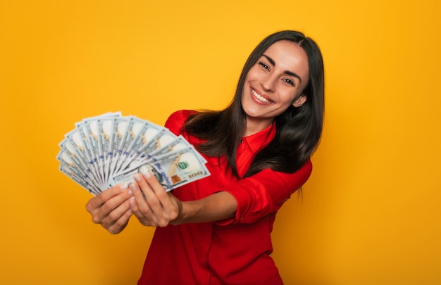 Young happy excited beautiful woman emotionally looking in the camera with a lot of money in her hands and having fun isolated on yellow background