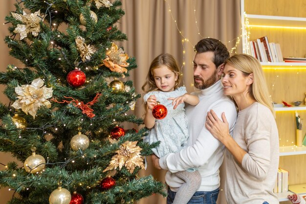 若い幸せな家族がクリスマス休暇のためにクリスマス ツリーを飾ります。お父さんは娘を中に抱えています。