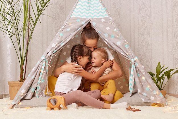 Young happy family in teepee tent loving mother with two kids sitting together in wigwam mom hugging her preschool and toddler kids in play tent Motherhood and childcare