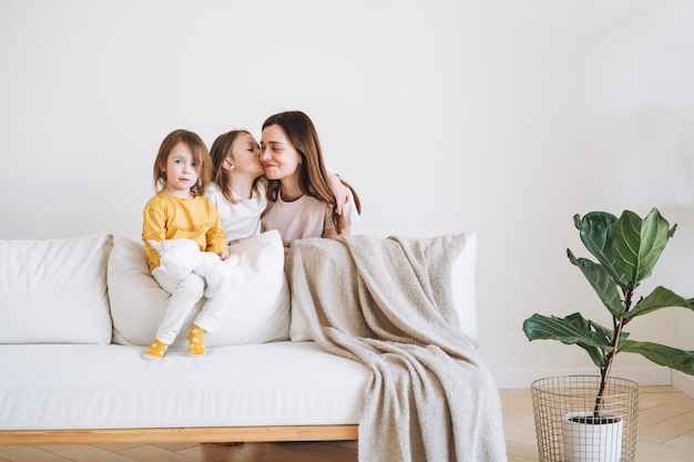 Young happy family with one parent woman mother with two children girls on couch in living room with green house plant at home