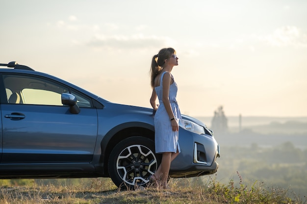 夏の自然の夕日の景色を楽しんで彼女の車の近くで休んでいる若い幸せな女性ドライバー。旅行の目的地とレクリエーションのコンセプト。