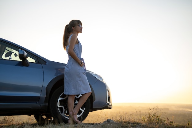 Молодая счастливая женщина-водитель отдыхает возле своей машины, наслаждаясь закатом летней природы.
