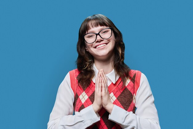 Photo young happy female in glasses pleading holding her palms together on blue studio background girl student looking at camera in request in plea asking for forgiveness for help sorry pray please