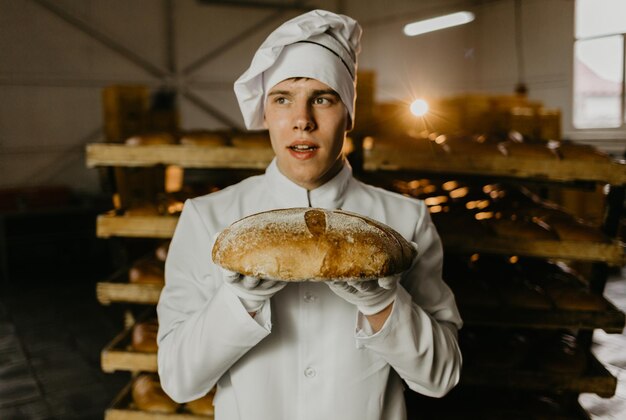 Giovane lavoratrice felice in panni sterili che tengono biscotti appena sfornati su banda stagnata all'interno della fabbrica di produzione alimentare