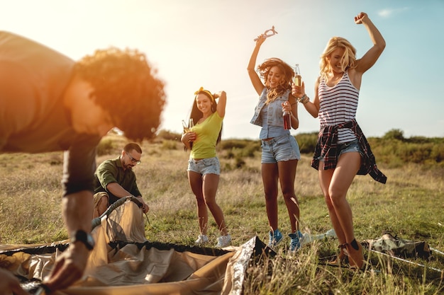 The young happy friends are preparing for camping. They're installing a tent on a suitable place in a meadow and their girlfriends are offering the beer to them. Girls dance in nature.