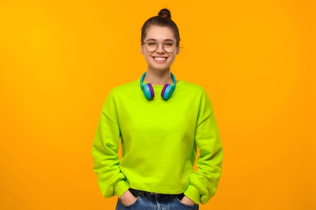 Photo young happy girl in green neon sweatshirt and jeans wearing wireless headphones around neck keeping hands in pockets isolated on yellow background
