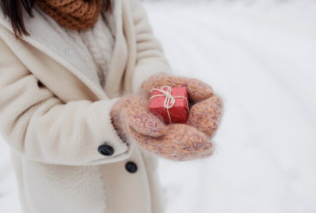 A young happy girl holds a gift box in knitted mittens in a snow-covered forest. Valentine's Day.