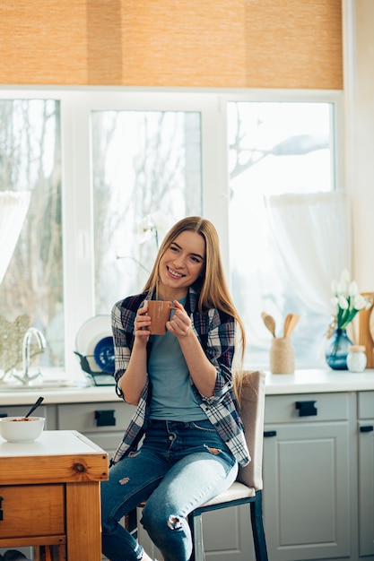 写真 彼女の手にマグカップを持ってキッチンに座って笑っている若い幸せな女の子