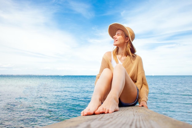 Young happy girl sitting on the pier barefoot and dreaming woman in a sun hat resting