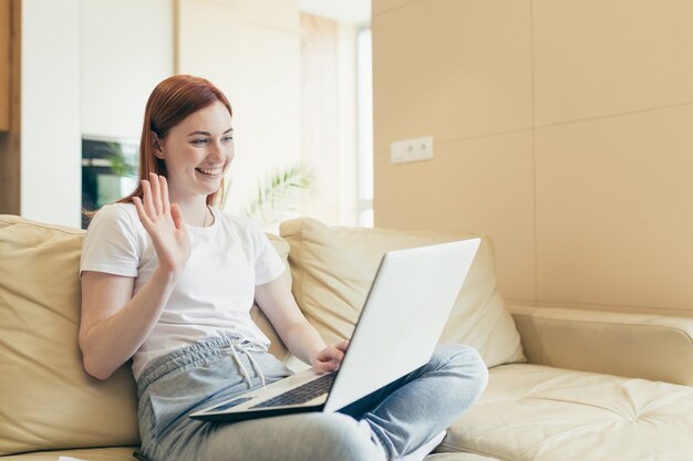 Young happy girl talking on video call online with laptop webcam at home showing heart gesture. Caucasian red haired woman sitting on couch communicates remotely with a loved one Love sincere emotions