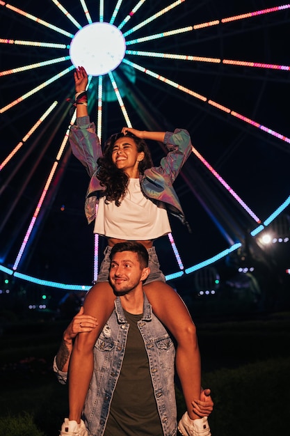 Young happy girlfriend sitting on the shoulders of her boyfriend and raising hand up in the amusement park