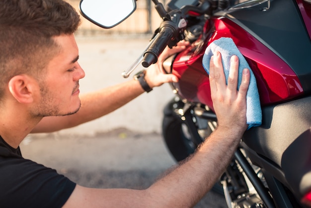 世話をし、屋外でバイクを掃除している若い幸せな男。ルビー色のオートバイを青い布で乾かします。