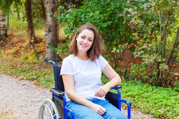 Young happy handicap woman in wheelchair on road in hospital park waiting for patient services. Paralyzed girl in invalid chair for disabled people outdoor in nature. Rehabilitation concept.