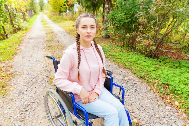 Young happy handicap woman in wheelchair on road in hospital park waiting for patient services. Paralyzed girl in invalid chair for disabled people outdoor in nature. Rehabilitation concept.