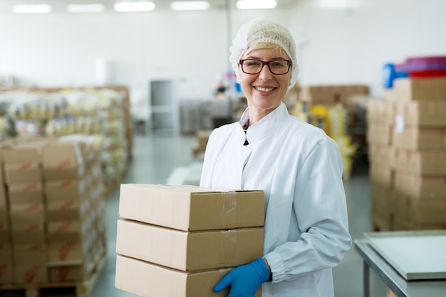 Young happy hard working female worker carrying a stack of boxes in factory storage room.