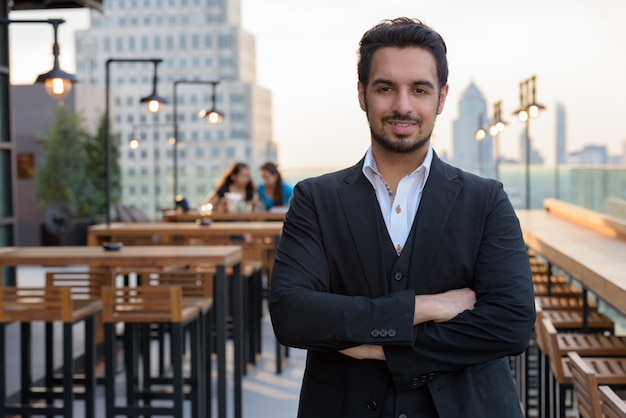 Young happy Indian businessman smiling with arms crossed at rooftop restaurant
