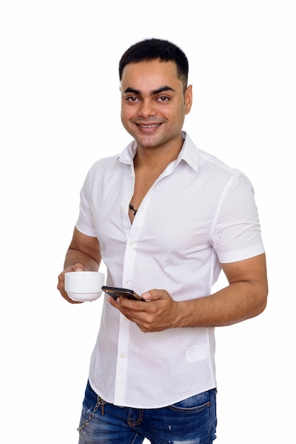 Giovane uomo indiano felice utilizzando il telefono cellulare mentre si tiene la tazza di caffè