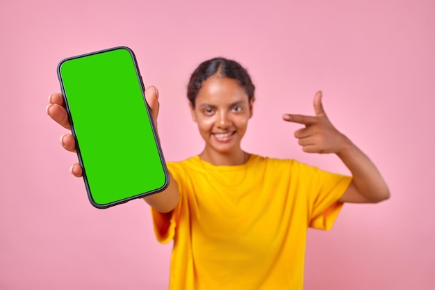 スタジオに立つ緑色の画面の携帯電話を示す若い幸せなインド人女性
