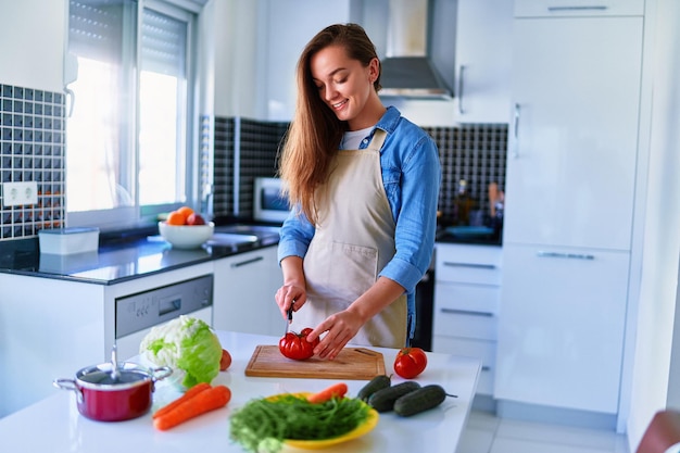 Young happy joyful cute smiling housewife preparing and cutting ripe vegetables on wooden board for fresh vegetarian salad for healthy eating