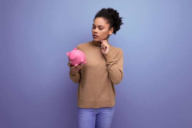 Young happy latin woman with curly hair holding piggy bank and isolated studio background with copy