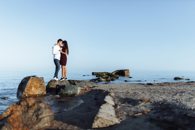 Young, happy, loving couple standing on the rocks at sea, in the arms, and looking at each other