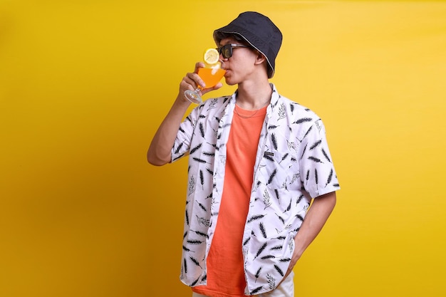 Young happy man in bucket hat and sunglasses drinking orange juice cocktail isolated on yellow studi