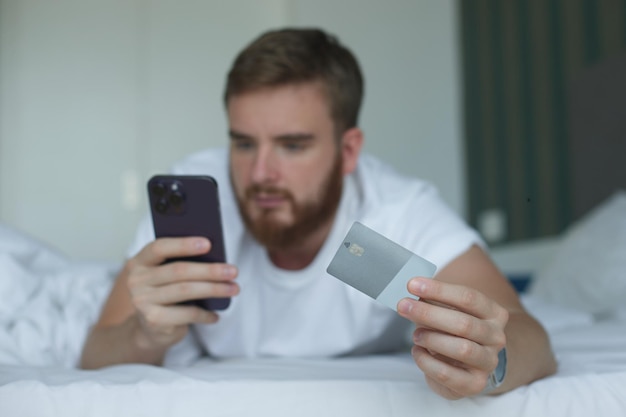 Young happy man doing online shopping with smartphone lay in bed in bedroom at home hold in hand