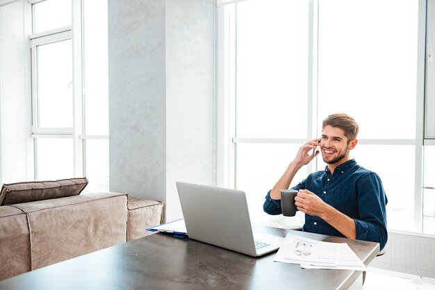 Young happy man drinking tea and sitting near table with laptop and documents while talking at his phone. Looking at laptop