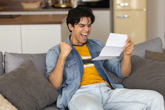 Young happy man holding a letter in his hands, reads the received notification with good news sitting on the couch at home