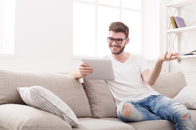 Young happy man at home having video call on tablet. Chatting with friend online on beige couch in light livingroom, copy space