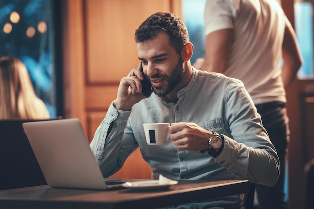 젊은 행복한 남자가 카페에서 전화를 받고 있습니다. 그는 고객과 이야기하고 노트북을보고 커피를 마시고 있습니다.
