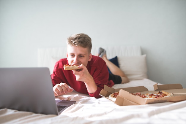 若い幸せな男はピザの箱が付いているベッドに横たわっています。