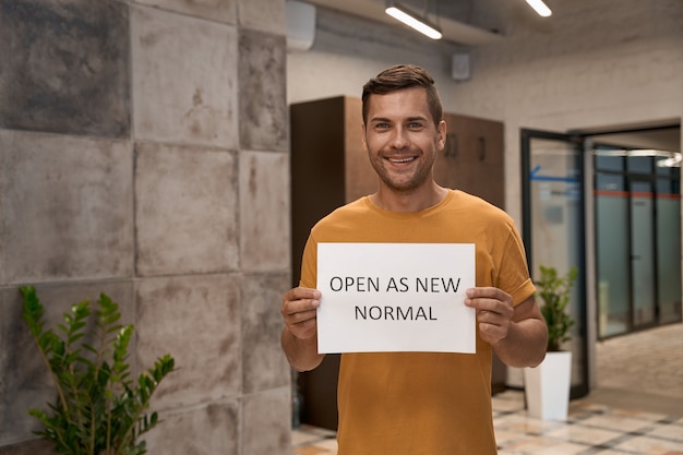 Young happy man office worker showing paper with text open as new normal at camera and smiling