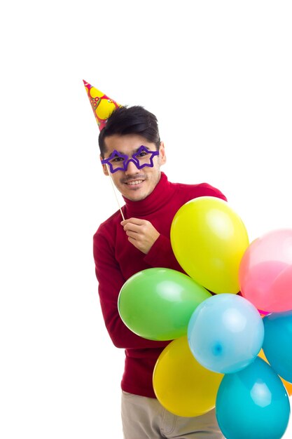 Giovane uomo felice in camicia rossa con cappello celebrativo che tiene molti palloncini colorati e carta stick di occhiali su sfondo bianco in studio