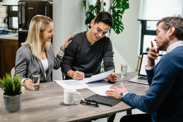 Young happy man signing mortgage contract while being with his wife on a meeting with real estate agent