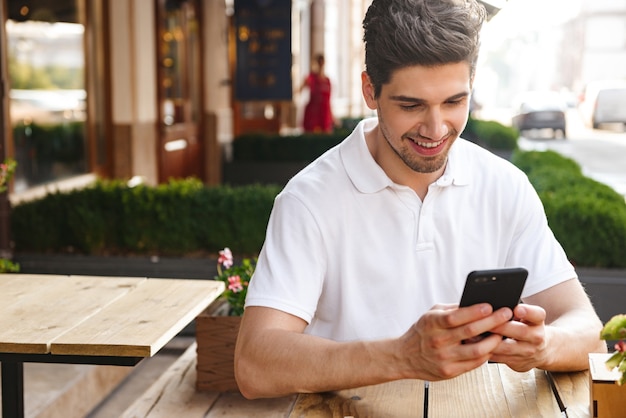 young happy man sitting in cafe outdoors while using mobile phone