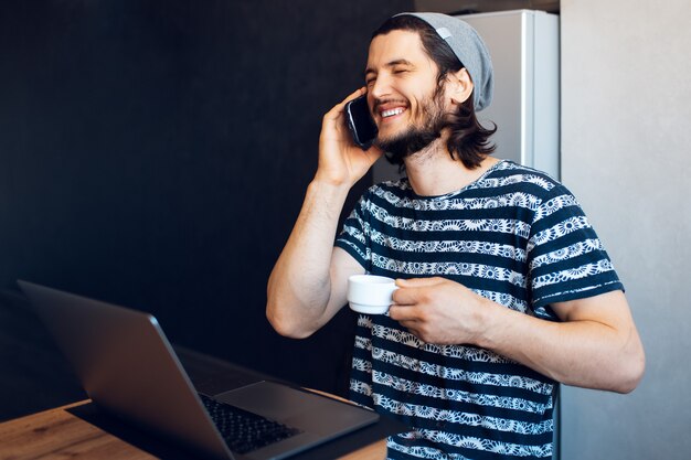 彼の電話で話し、彼のラップトップで作業し、一杯のコーヒーを持っている若い幸せな男