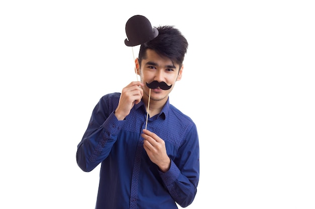 Young happy man with dark hair in blue shirt holding cardboard sticks of moustache and black hat on white background in studio