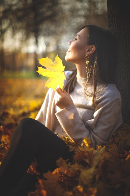 Young happy mixed race woman sitting near tree in autumn park looking at sunset