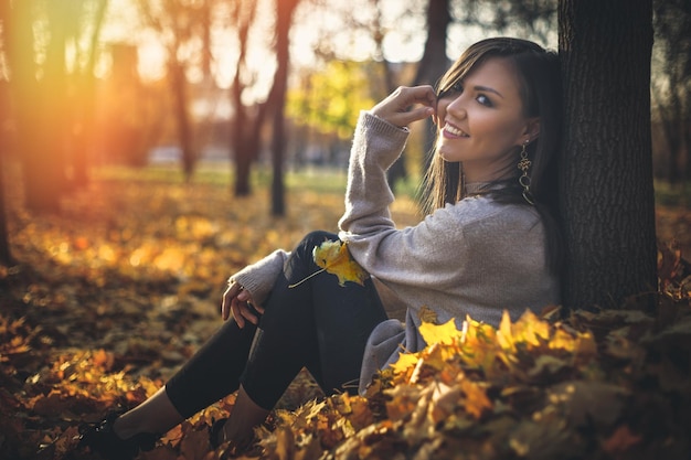 Young happy mixed race woman sitting near tree in autumn park at sunset looking at camera