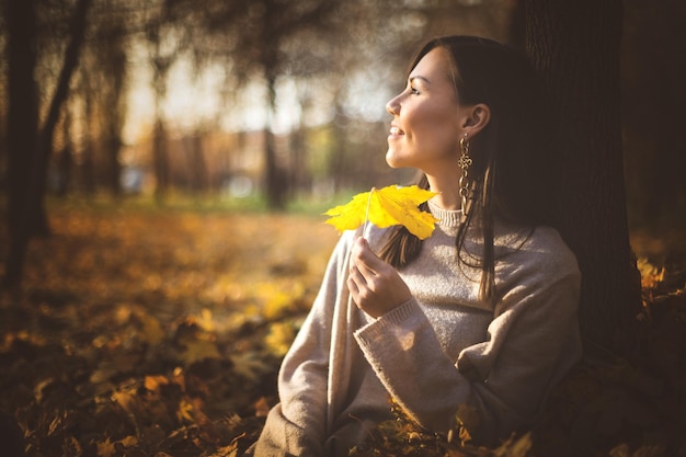 Giovane donna felice di razza mista seduta vicino all'albero nel parco autunnale al tramonto